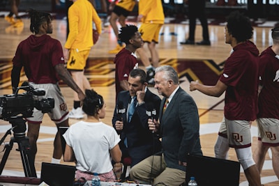 A group of individuals, likely basketball players, is on a gymnasium court. Some are wearing yellow athletic outfits, and others are in burgundy athletic gear. Two men in suits are speaking into microphones while a woman stands nearby wearing a white shirt. A camera setup is positioned to capture the scene, indicating a broadcast or interview setting.