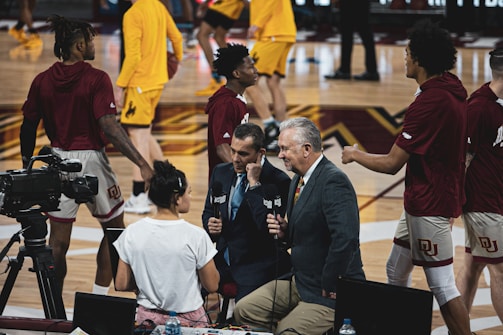 A group of individuals, likely basketball players, is on a gymnasium court. Some are wearing yellow athletic outfits, and others are in burgundy athletic gear. Two men in suits are speaking into microphones while a woman stands nearby wearing a white shirt. A camera setup is positioned to capture the scene, indicating a broadcast or interview setting.