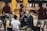 A group of individuals, likely basketball players, is on a gymnasium court. Some are wearing yellow athletic outfits, and others are in burgundy athletic gear. Two men in suits are speaking into microphones while a woman stands nearby wearing a white shirt. A camera setup is positioned to capture the scene, indicating a broadcast or interview setting.