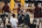 A group of individuals, likely basketball players, is on a gymnasium court. Some are wearing yellow athletic outfits, and others are in burgundy athletic gear. Two men in suits are speaking into microphones while a woman stands nearby wearing a white shirt. A camera setup is positioned to capture the scene, indicating a broadcast or interview setting.