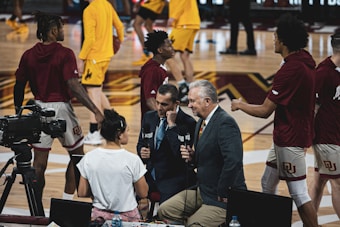 A group of individuals, likely basketball players, is on a gymnasium court. Some are wearing yellow athletic outfits, and others are in burgundy athletic gear. Two men in suits are speaking into microphones while a woman stands nearby wearing a white shirt. A camera setup is positioned to capture the scene, indicating a broadcast or interview setting.