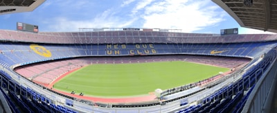 A large, empty football stadium with a green field and surrounding tiers of seats. The stands are decorated with designs, including large yellow and blue banners. The sky is partly cloudy, adding a bright ambiance. The phrase 'Més que un club' is prominently displayed across one of the seating tiers.
