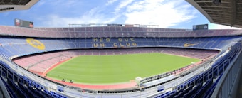 A large, empty football stadium with a green field and surrounding tiers of seats. The stands are decorated with designs, including large yellow and blue banners. The sky is partly cloudy, adding a bright ambiance. The phrase 'M&eacute;s que un club' is prominently displayed across one of the seating tiers.