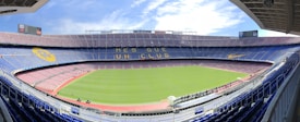 A large, empty football stadium with a green field and surrounding tiers of seats. The stands are decorated with designs, including large yellow and blue banners. The sky is partly cloudy, adding a bright ambiance. The phrase 'M&eacute;s que un club' is prominently displayed across one of the seating tiers.