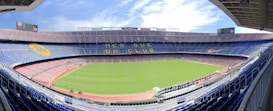 A large, empty football stadium with a green field and surrounding tiers of seats. The stands are decorated with designs, including large yellow and blue banners. The sky is partly cloudy, adding a bright ambiance. The phrase 'M&eacute;s que un club' is prominently displayed across one of the seating tiers.