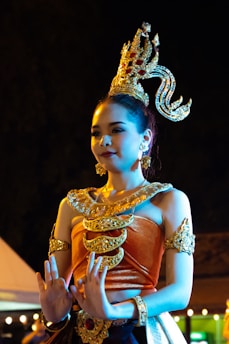 A person wearing traditional ornate attire with gold embellishments and an intricate headdress stands with hands raised gracefully. The lighting creates a dramatic and colorful effect, highlighting the rich textures and details of the costume.
