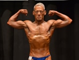 A muscular individual is flexing their arms on stage, showcasing strong definition and muscularity. They are wearing a blue posing suit and standing against a dark background.