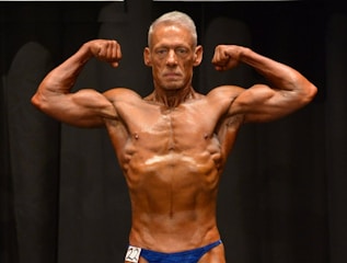 A muscular individual is flexing their arms on stage, showcasing strong definition and muscularity. They are wearing a blue posing suit and standing against a dark background.