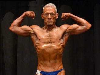 A muscular individual is flexing their arms on stage, showcasing strong definition and muscularity. They are wearing a blue posing suit and standing against a dark background.