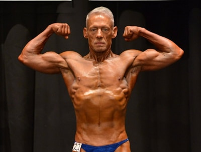 A muscular individual is flexing their arms on stage, showcasing strong definition and muscularity. They are wearing a blue posing suit and standing against a dark background.