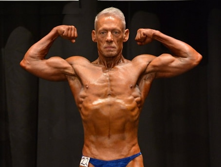 A muscular individual is flexing their arms on stage, showcasing strong definition and muscularity. They are wearing a blue posing suit and standing against a dark background.