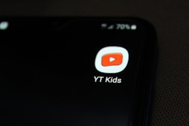 A smartphone screen displaying the YT Kids app icon, which features a red play button inside a white rounded square. The icon is prominently displayed against a dark background, with a battery icon at the top right corner showing 70% charge.