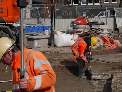 Construction workers in orange reflective safety gear are actively working on a building site. One worker is drilling into the ground, while another holds a survey pole. The area is cluttered with tools, concrete, wood pallets, and equipment such as a cement mixer. A temporary fence surrounds the site.