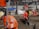 Construction workers in orange reflective safety gear are actively working on a building site. One worker is drilling into the ground, while another holds a survey pole. The area is cluttered with tools, concrete, wood pallets, and equipment such as a cement mixer. A temporary fence surrounds the site.