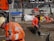 Construction workers in orange reflective safety gear are actively working on a building site. One worker is drilling into the ground, while another holds a survey pole. The area is cluttered with tools, concrete, wood pallets, and equipment such as a cement mixer. A temporary fence surrounds the site.