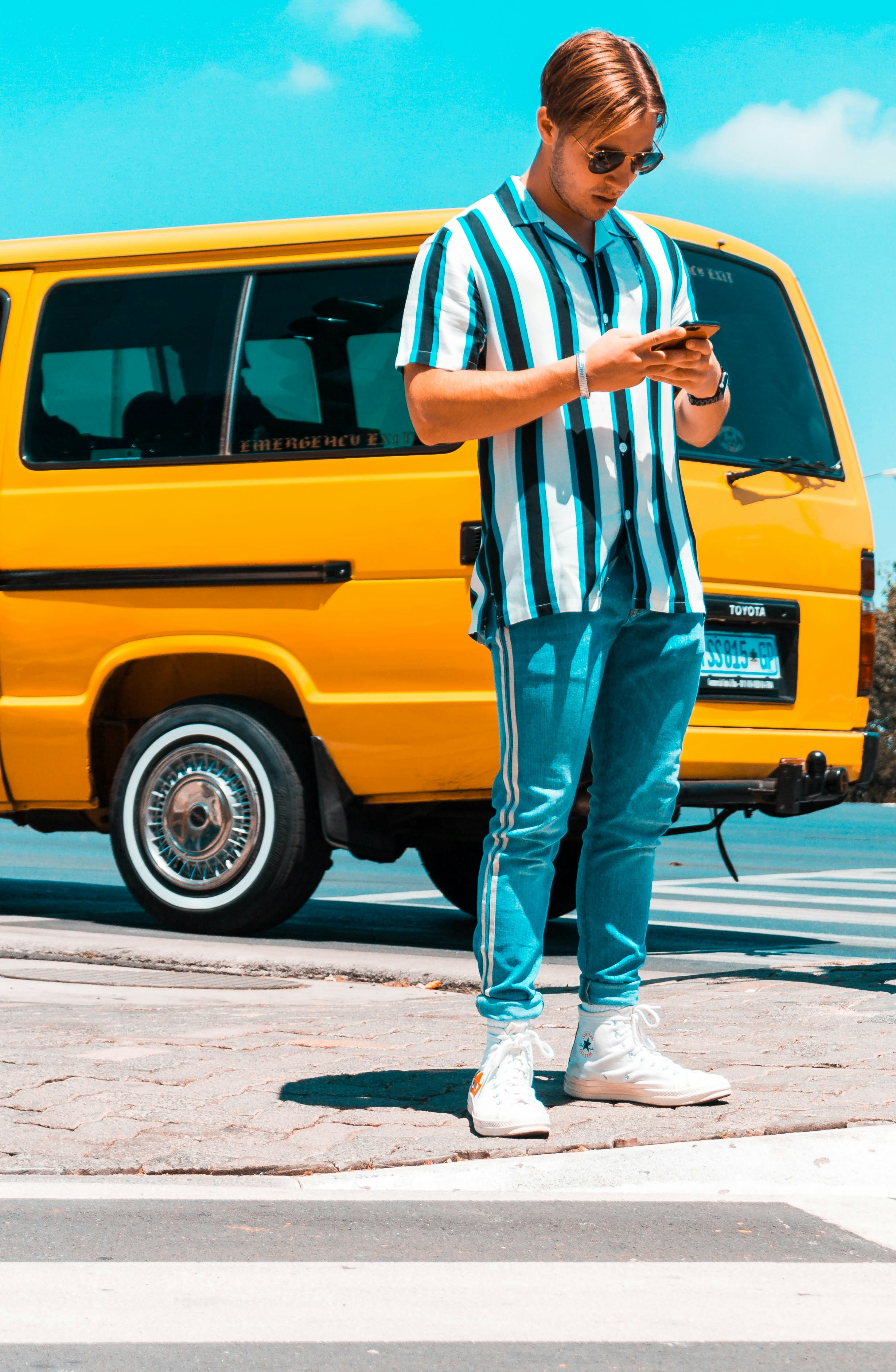 Pria mengenakan kemeja bergaris putih dan hitam dan celana biru menggunakan smartphone berdiri di samping van kuning di siang hari