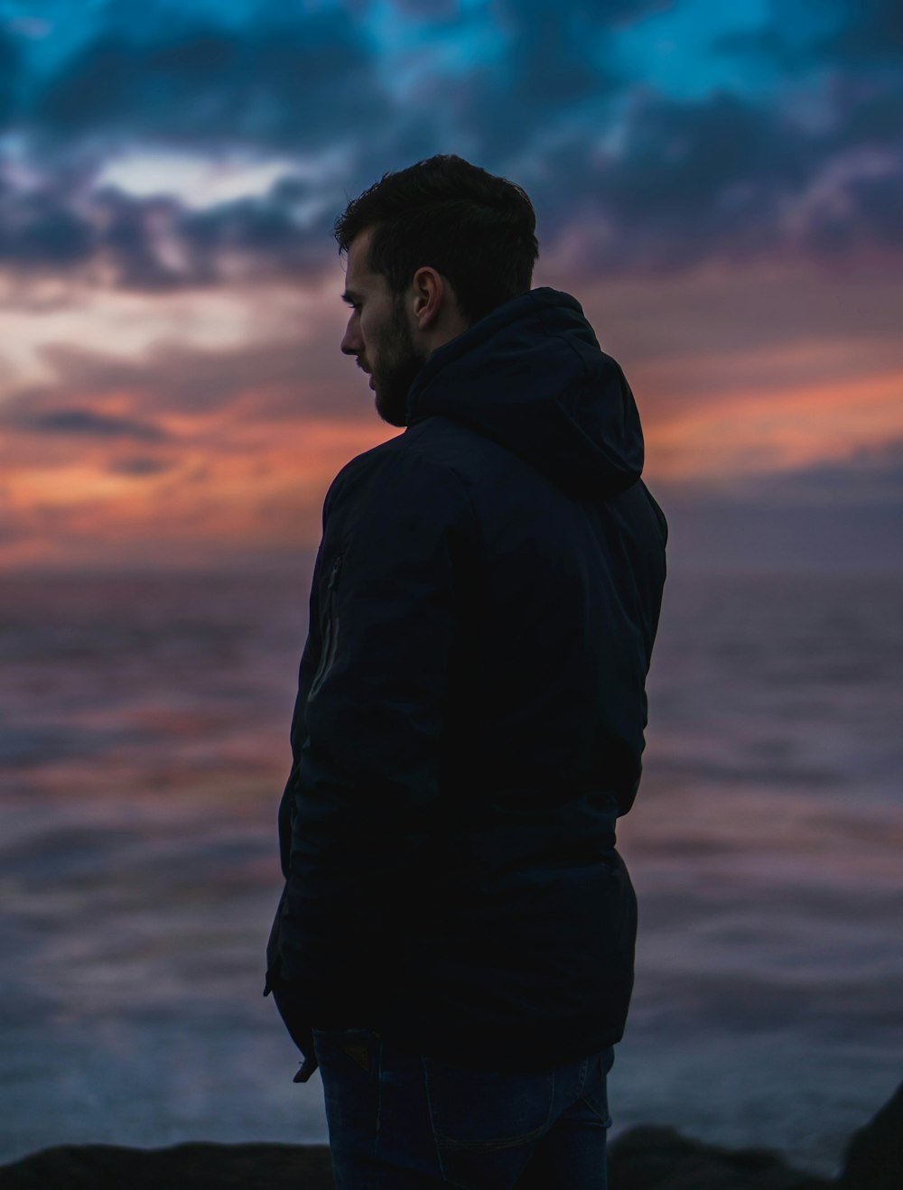 man wearing black jacket standing while facing his right side under blue and orange sky