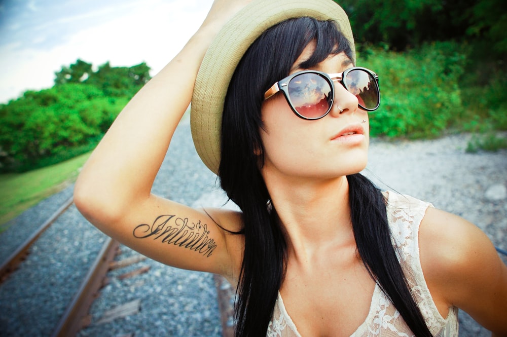 portrait photography of woman wearing white floral sleeveless blouse, sunglasses, and white hat glancing her left side while touching hat