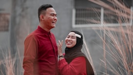 A couple laughing and enjoying each other's company. Both are dressed in red, with one wearing a black hijab and displaying a white hair clip. They are surrounded by tall grassy plants with a blurred background.