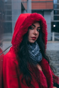 A woman wearing a bright red hooded coat stands against a metallic surface with an urban background. Her long dark hair flows over her shoulders, and she has a pensive expression. Around her neck is a thick, dark scarf.