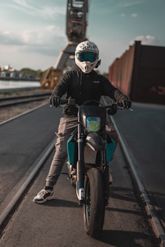 A person wearing a white helmet and protective jacket is sitting on a motorcycle on a paved road. Behind them, an industrial crane and rails can be seen, with a river and some buildings in the background. The lighting suggests a late afternoon setting.