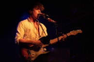 A musician with curly hair is performing on stage. The person is wearing a light-colored, patterned shirt and is playing an electric guitar. The stage is dimly lit with a spotlight focused on the musician, creating an intimate atmosphere.