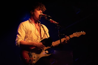 A musician with curly hair is performing on stage. The person is wearing a light-colored, patterned shirt and is playing an electric guitar. The stage is dimly lit with a spotlight focused on the musician, creating an intimate atmosphere.