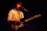 A musician with curly hair is performing on stage. The person is wearing a light-colored, patterned shirt and is playing an electric guitar. The stage is dimly lit with a spotlight focused on the musician, creating an intimate atmosphere.