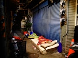A person lies on a mattress in a dimly lit alleyway. The area is cluttered with objects, including a bicycle basket holding a plastic bottle, a red umbrella, and traffic cones. The alley features a blue metal shutter and several plants in pots, with infrastructure like pipes and utility meters visible along the wall.