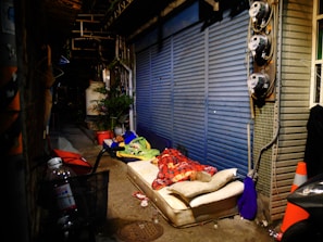 A person lies on a mattress in a dimly lit alleyway. The area is cluttered with objects, including a bicycle basket holding a plastic bottle, a red umbrella, and traffic cones. The alley features a blue metal shutter and several plants in pots, with infrastructure like pipes and utility meters visible along the wall.