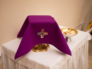 A purple cloth with a small embroidered cross covers a sacred item on an altar. Next to it, a silver platter holds several round pieces of unleavened bread. The altar is draped with a white cloth, set against a plain beige wall.