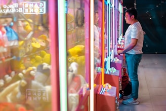 A person wearing a striped shirt stands in front of a series of brightly lit claw machines filled with various plush toys. The scene is illuminated with vibrant neon lights, casting colorful reflections on the glass surfaces.
