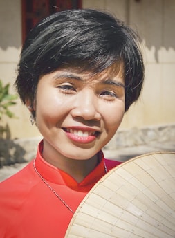 A person smiles while holding a traditional Asian bamboo hat, wearing a bright red outfit with a necklace. The background includes a soft-focus exterior setting with a building.