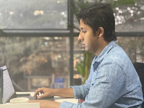 A person wearing a blue shirt is sitting at a desk and working on a laptop. There is a window in the background, and the setting appears to be a modern office with natural light filtering through the large glass panes. A notebook and a cup are on the desk, indicating a work or study environment.