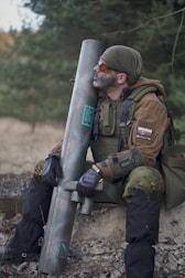 A person dressed in military gear sits outdoors holding a large cylindrical object. The setting is natural, with trees and grass in the background. The individual wears camouflage gloves, a tactical vest, and protective goggles. Earthy tones dominate the attire, suggesting a tactical or military scenario.