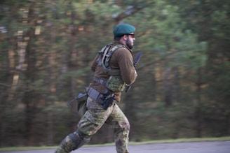 A person wearing camouflage clothing and a green beret is running outdoors. The individual is carrying tactical gear and appears to be in motion on a blurred background of green trees.