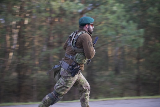 A person wearing camouflage clothing and a green beret is running outdoors. The individual is carrying tactical gear and appears to be in motion on a blurred background of green trees.