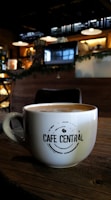A cozy cafe environment with a large white coffee mug placed on a wooden table. The mug features the phrase 'Cafe Central' in a modern font. The background features dim lighting, with hanging lamps, a menu board, and coffee machines creating an inviting atmosphere.