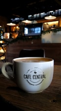 A cozy cafe environment with a large white coffee mug placed on a wooden table. The mug features the phrase 'Cafe Central' in a modern font. The background features dim lighting, with hanging lamps, a menu board, and coffee machines creating an inviting atmosphere.