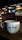 A cozy cafe environment with a large white coffee mug placed on a wooden table. The mug features the phrase 'Cafe Central' in a modern font. The background features dim lighting, with hanging lamps, a menu board, and coffee machines creating an inviting atmosphere.