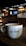 A cozy cafe environment with a large white coffee mug placed on a wooden table. The mug features the phrase 'Cafe Central' in a modern font. The background features dim lighting, with hanging lamps, a menu board, and coffee machines creating an inviting atmosphere.
