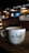 A cozy cafe environment with a large white coffee mug placed on a wooden table. The mug features the phrase 'Cafe Central' in a modern font. The background features dim lighting, with hanging lamps, a menu board, and coffee machines creating an inviting atmosphere.