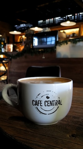 A cozy cafe environment with a large white coffee mug placed on a wooden table. The mug features the phrase 'Cafe Central' in a modern font. The background features dim lighting, with hanging lamps, a menu board, and coffee machines creating an inviting atmosphere.