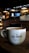 A cozy cafe environment with a large white coffee mug placed on a wooden table. The mug features the phrase 'Cafe Central' in a modern font. The background features dim lighting, with hanging lamps, a menu board, and coffee machines creating an inviting atmosphere.