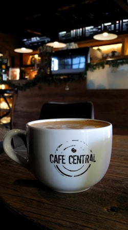 A cozy cafe environment with a large white coffee mug placed on a wooden table. The mug features the phrase 'Cafe Central' in a modern font. The background features dim lighting, with hanging lamps, a menu board, and coffee machines creating an inviting atmosphere.