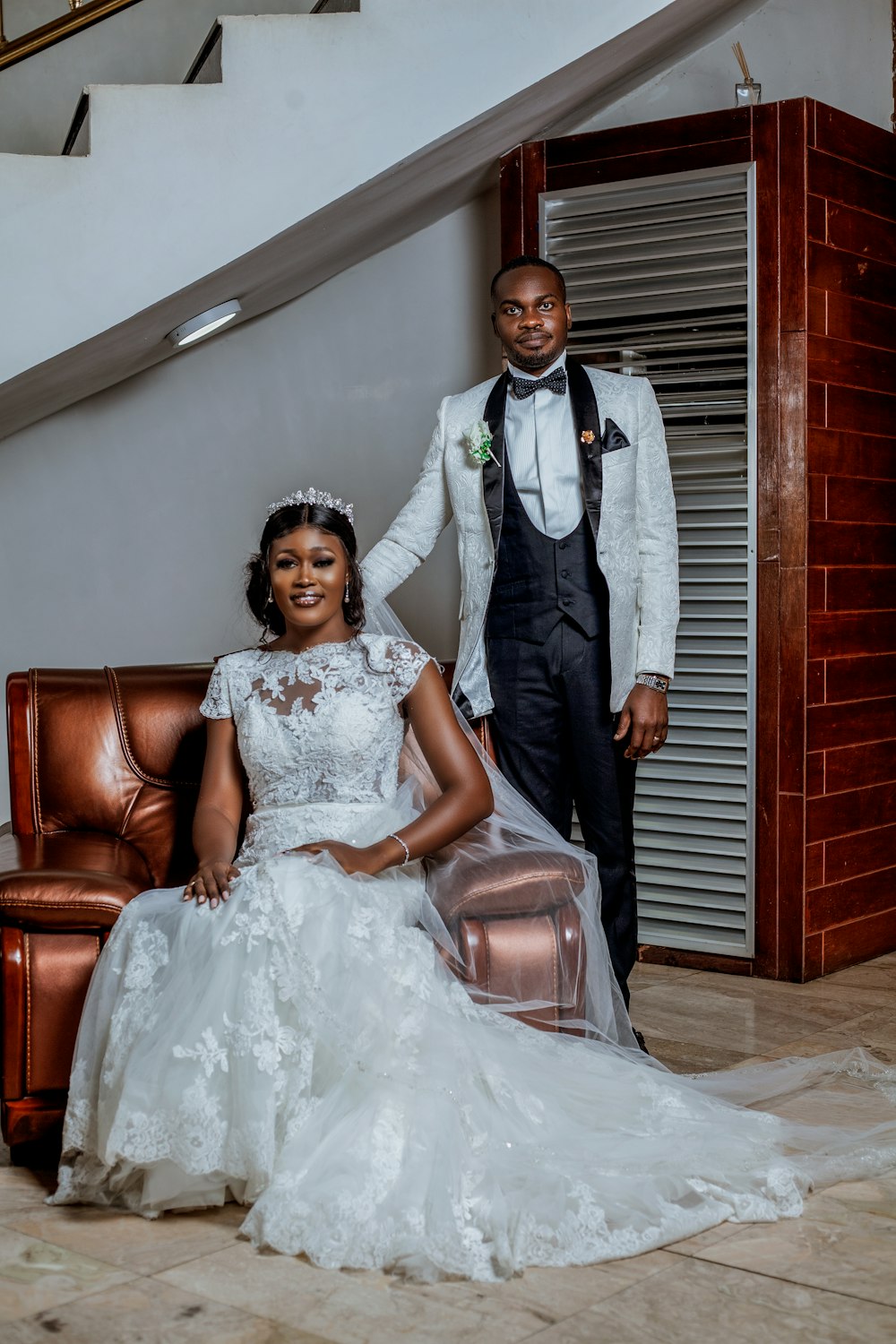 groom standing beside a bride sitting on a sofa chair