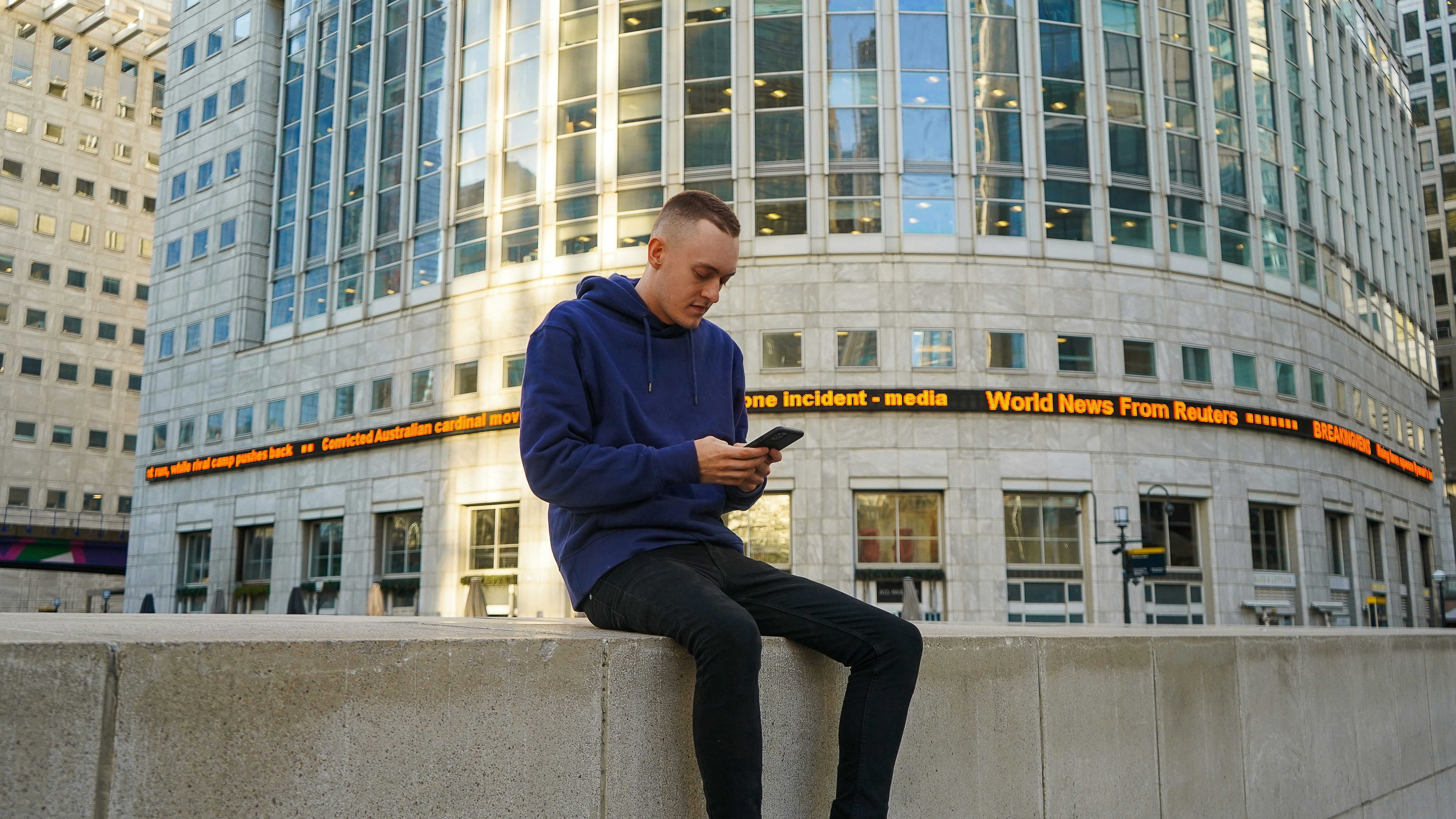 James Frewin (British Designer) using his Phone while sitting on a wall outside Canary Wharf in London, United Kingdom.