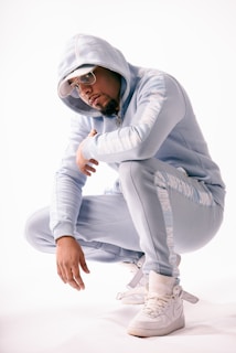 A person is crouching while wearing a light blue tracksuit with white lettering on the sleeves and legs. They have on a hood, sunglasses, and a white cap, paired with white high-top sneakers. The background is plain and brightly lit, giving a clean and modern look.