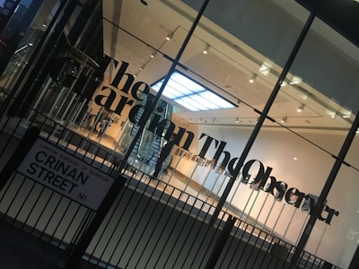 A modern glass building entrance with large black lettering spelling out a newspaper name on the glass. The interior appears well-lit with a visible staircase, minimalistic design, and a sign with a street name in the foreground.