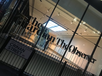 A modern glass building entrance with large black lettering spelling out a newspaper name on the glass. The interior appears well-lit with a visible staircase, minimalistic design, and a sign with a street name in the foreground.